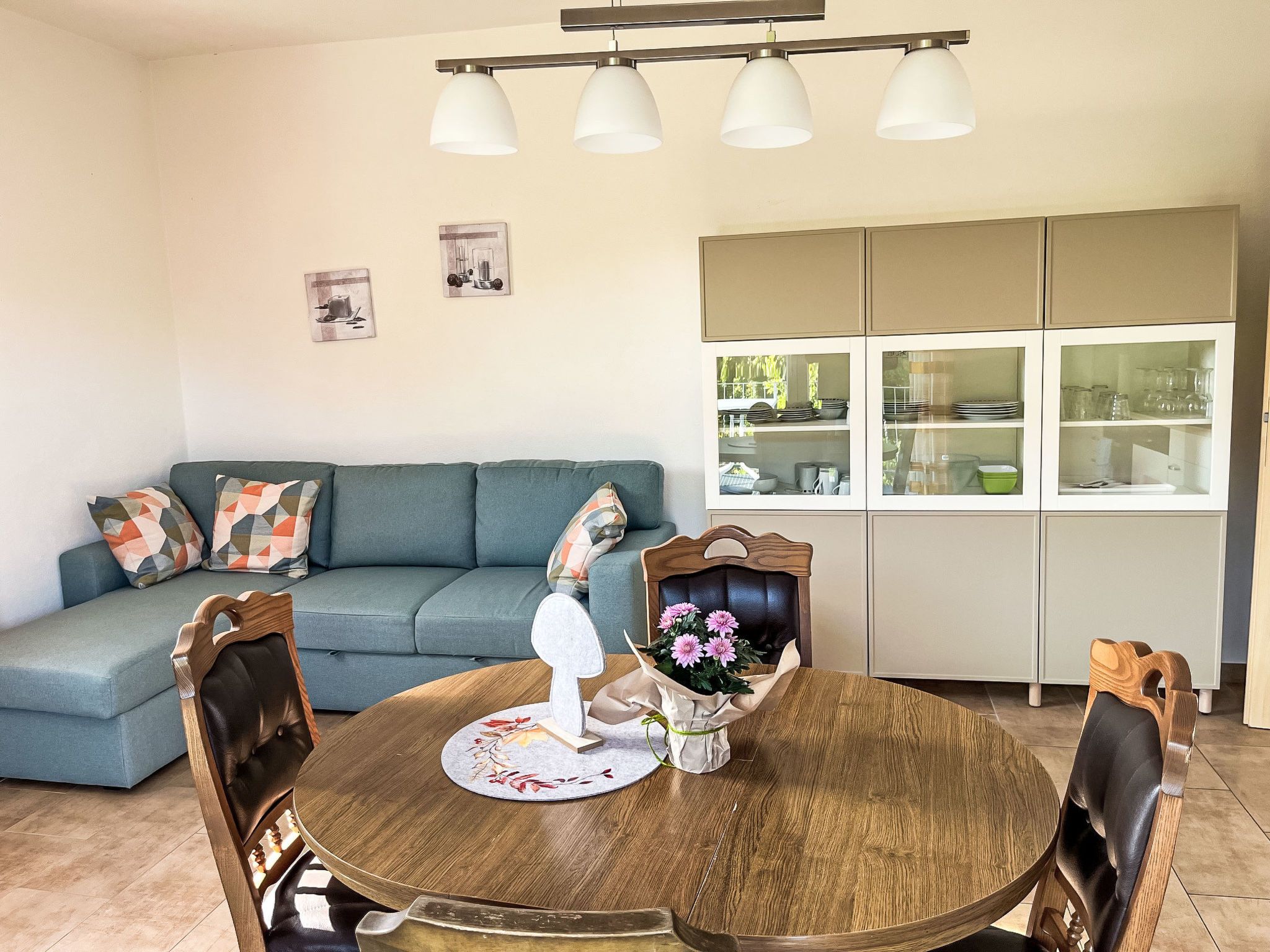 Ein Wohnzimmer mit blauem Sofa, einem Holzesstisch mit Stühlen und einer Küchenzeile mit Schränken und Gläsern.