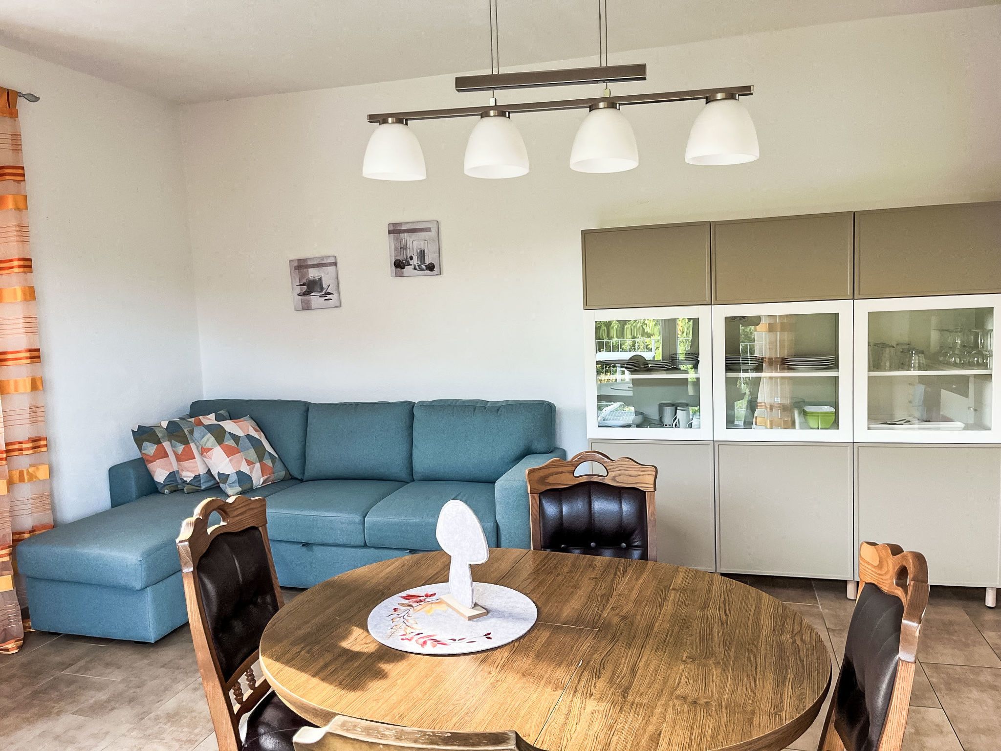 A modern living room with a blue sofa, a wooden dining table with chairs, and a kitchen area with cabinets and a window.