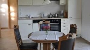 Un comedor con una mesa redonda y cuatro sillas, una cocina con muebles blancos y una estufa, y un colgador de luz.