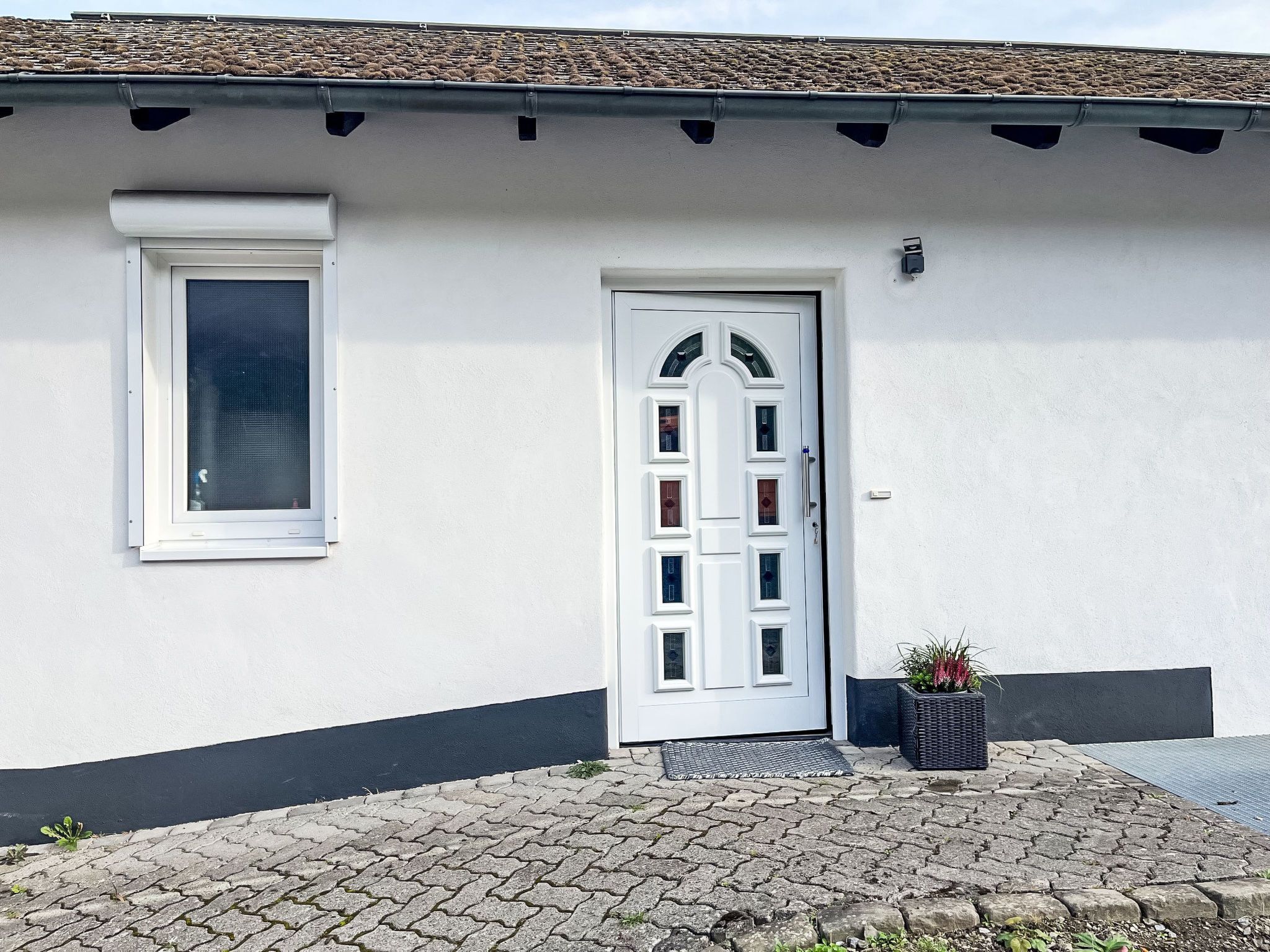 Ein weißes Gebäude mit grauem Dach, einer weißen Tür mit Glasscheiben, einem Fenster mit weißem Rahmen und einem kleinen Topfpflanzen neben der Tür.