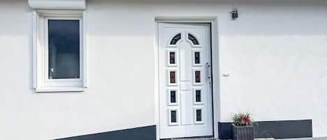 A white building with a grey roof, a white door with glass panels, a window with a white frame, and a small potted plant beside the door.