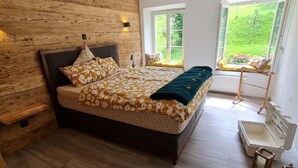 Ein Schlafzimmer mit einer Holzwand, einem Bett mit gemusterten Betttextilien, zwei Fenstern und einem Nachttisch mit einer Lampe.