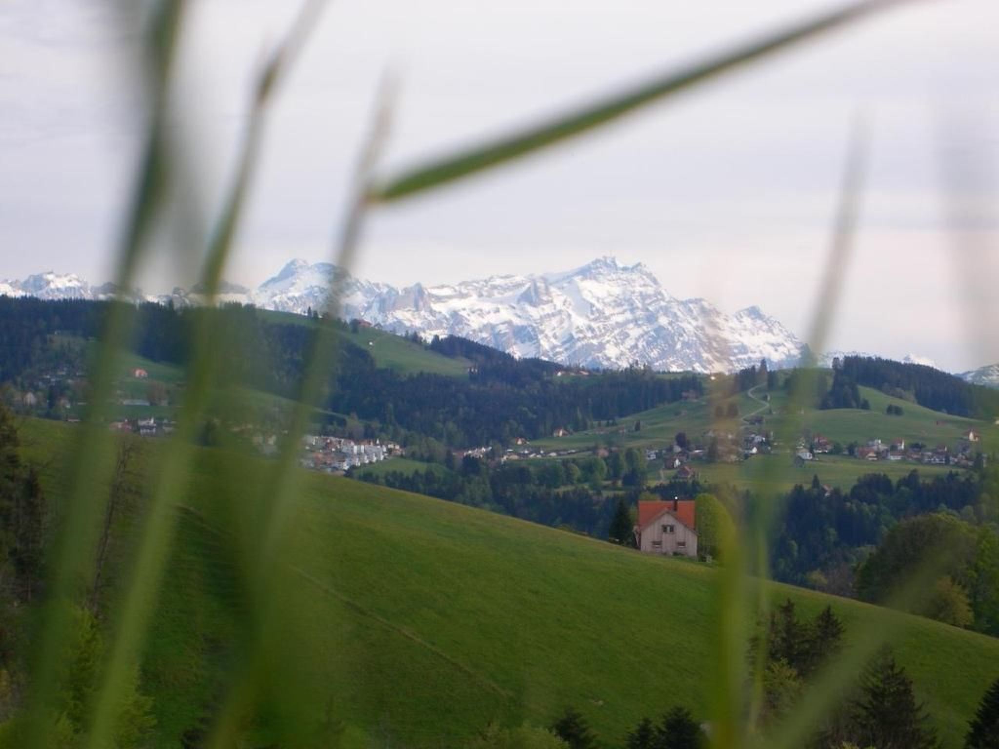 Paesaggio rurale con una casa su una collina, un villaggio sottostante e montagne innevate sullo sfondo.