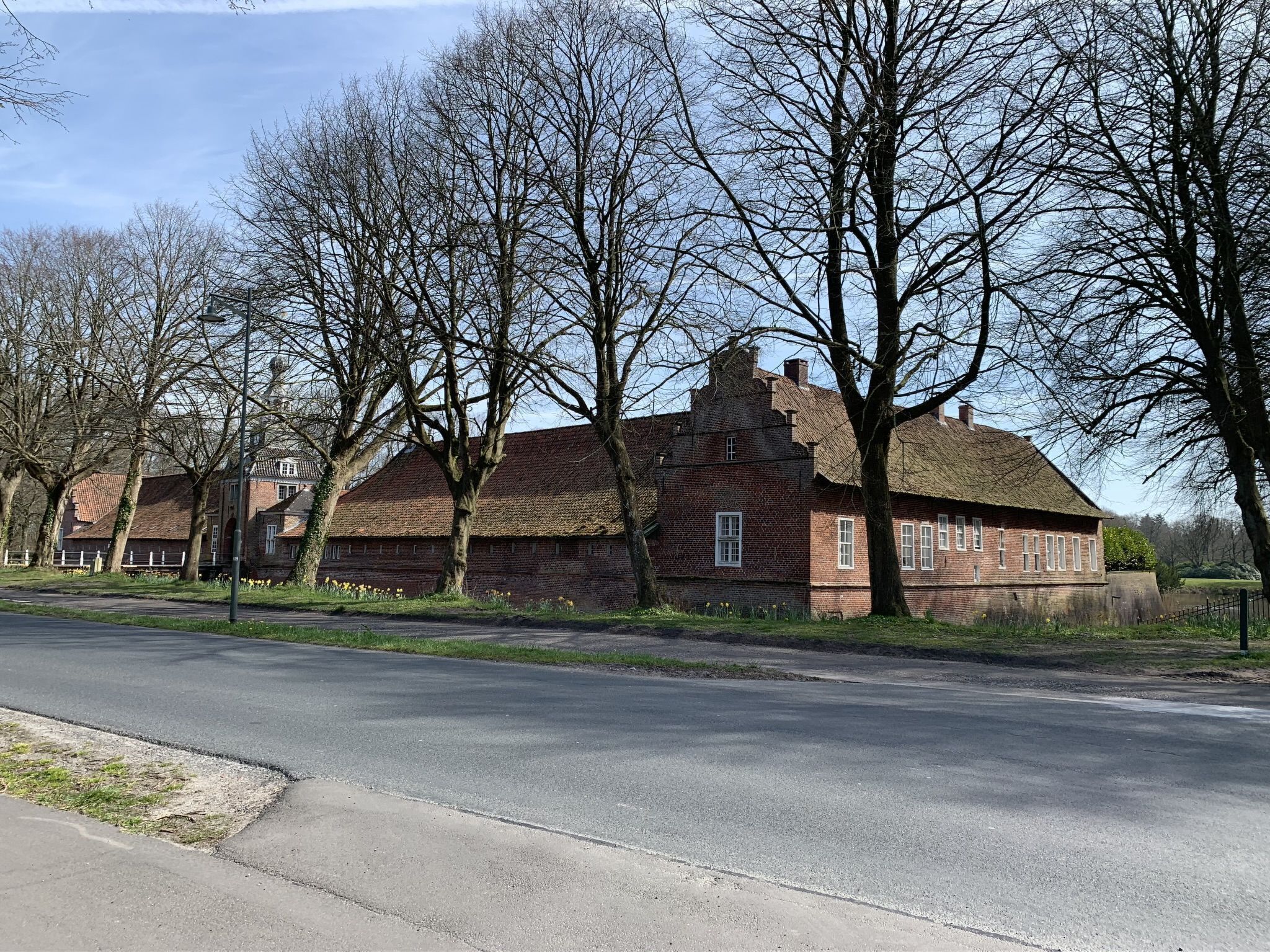 En traditionel murbygning med stråtækt tag, omgivet af blottede træer og en belagt vej.