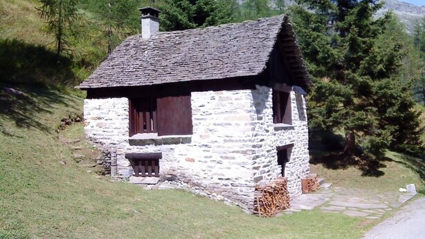Una pequeña cabaña de piedra con techo inclinado, contraventanas de madera y chimenea.