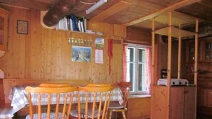 Interior de una cabaña de madera con una mesa de comedor, sillas, una ventana con cortinas y una zona de cocina.