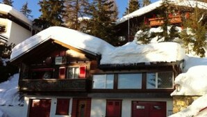 A two-story lodge with a snow-covered roof and red doors, surrounded by trees and snow. - Chalet Adler- Studio für 2 Personen by Interhome (Aminona)