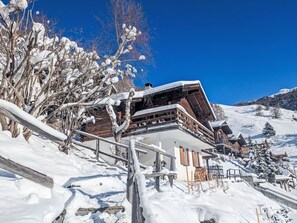 Un chalet en bois, doté d’un toit en pente, entouré d’arbres couverts de neige et surplombé d’un ciel bleu clair.