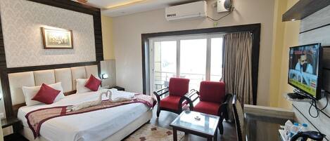 A hotel room with a bed, two red armchairs, a glass coffee table, a TV, and an air conditioning unit.