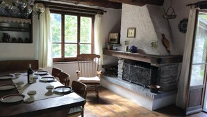 Un coin repas rustique avec une table en bois dressée pour quatre personnes, une cheminée en pierre et une fenêtre agrémentée de rideaux.