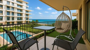 Uma varanda com uma cadeira suspensa, uma mesa e duas cadeiras com vista para uma piscina e um edifício de hotel.