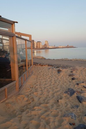 Blick auf den Strand mit einer hölzernen Struktur, vermutlich ein Restaurant, mit Blick auf das Meer und eine entfernte Skyline.