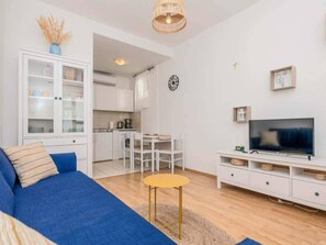 Ein modernes Wohnzimmer mit einem blauen Sofa, einem kleinen runden Tisch, einem Fernsehgerät auf einer weißen Kommode und einer Küche im Hintergrund.