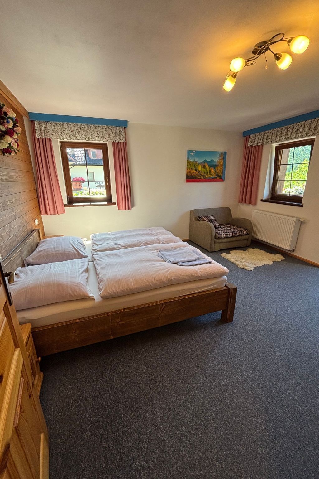 Un dormitorio con una cama de madera, dos ventanas con cortinas, una silla, un radiador y un cuadro en la pared.