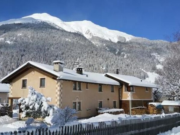 Un bâtiment à deux étages, dont le toit est recouvert de neige, entouré d’une clôture en bois et d’arbres également couverts de neige.