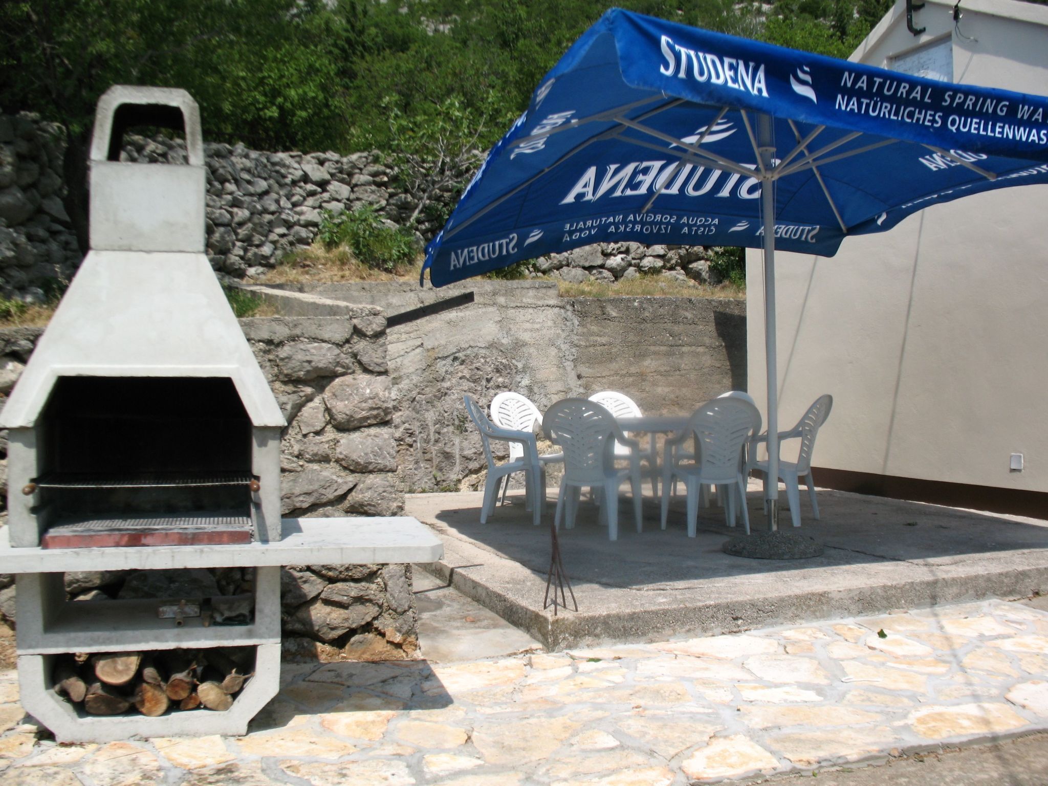 Area barbecue esterna con parete a muro in pietra, un ombrello blu con la scritta 'STUDENA' e un tavolo con sedie.