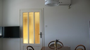 Spiseområde med et træbord, stole, et fladskærms-tv monteret på væggen og et vindue med persienner.