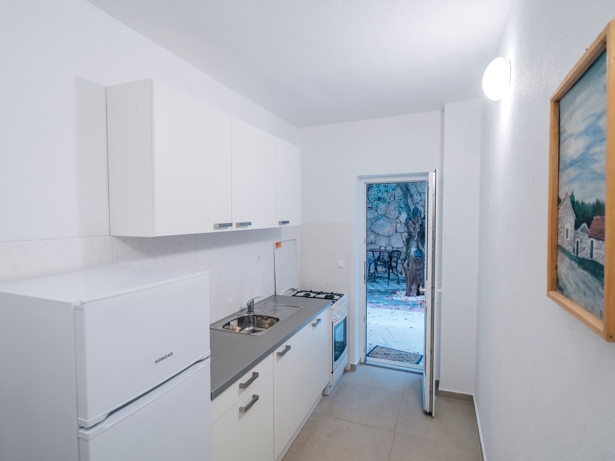 A compact kitchen with white cabinets, a gas stove, and a sink. A door leads to a small outdoor area with a stone wall and a chair.