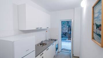 A compact kitchen with white cabinets, a gas stove, and a sink. A door leads to a small outdoor area with a stone wall and a chair.