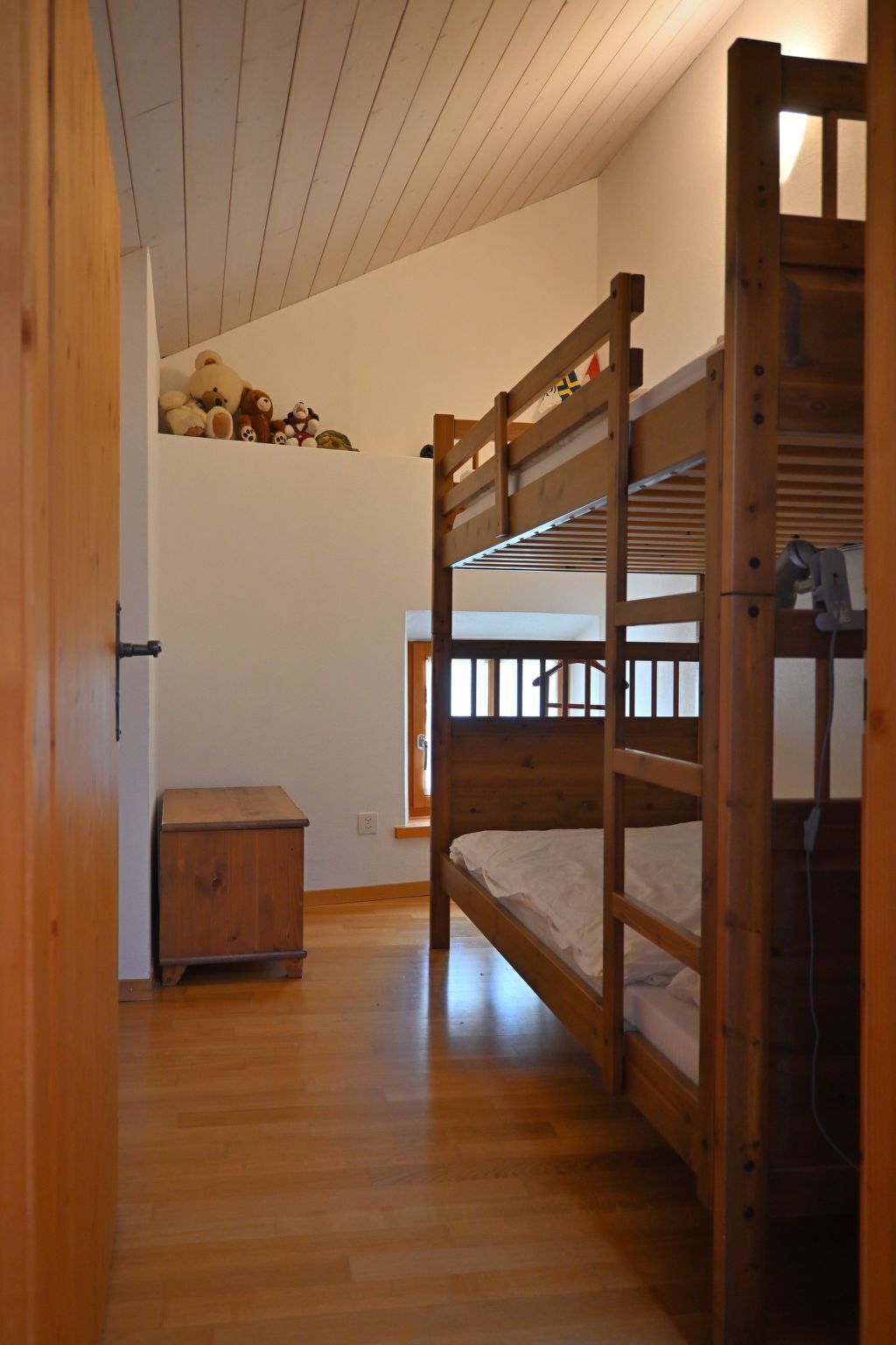 Habitación con literas, suelo de madera, un pequeño cofre de madera y una ventana.