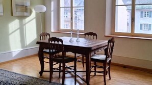 Un comedor con una mesa de madera y cuatro sillas, ventanas amplias con vista a edificios, y una lámpara de pie.