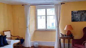 Una habitación con una pared amarilla, un sofá blanco con cojines azules, un sillón rojo, una mesita con una lámpara y una ventana con cortinas blancas.