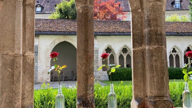 Un arco de piedra con vista despejada a un patio interior, que incluye un edificio con tejado rojo y una rosa en un frasco.