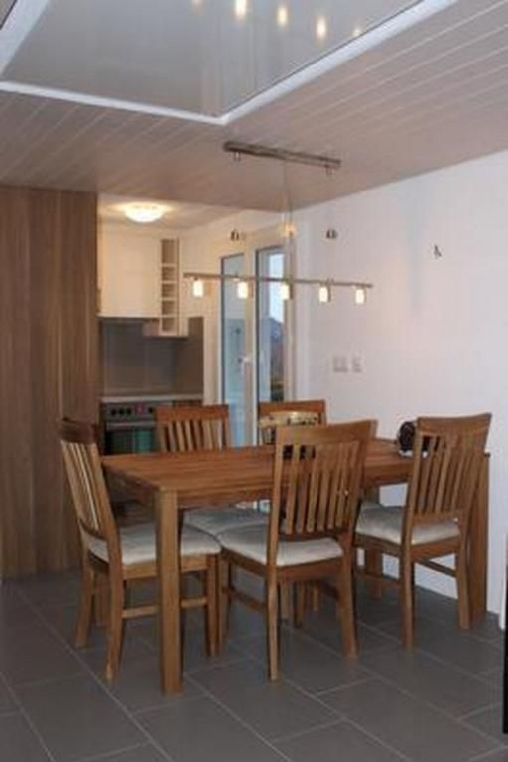 Un comedor con mesa y sillas de madera, una cocina al fondo y un techo con iluminación empotrada.