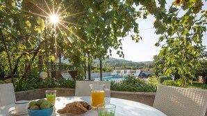 Et bord dækket udendørs med morgenmad, et glas juice og udsigt til en pool og grøn bevoksning.