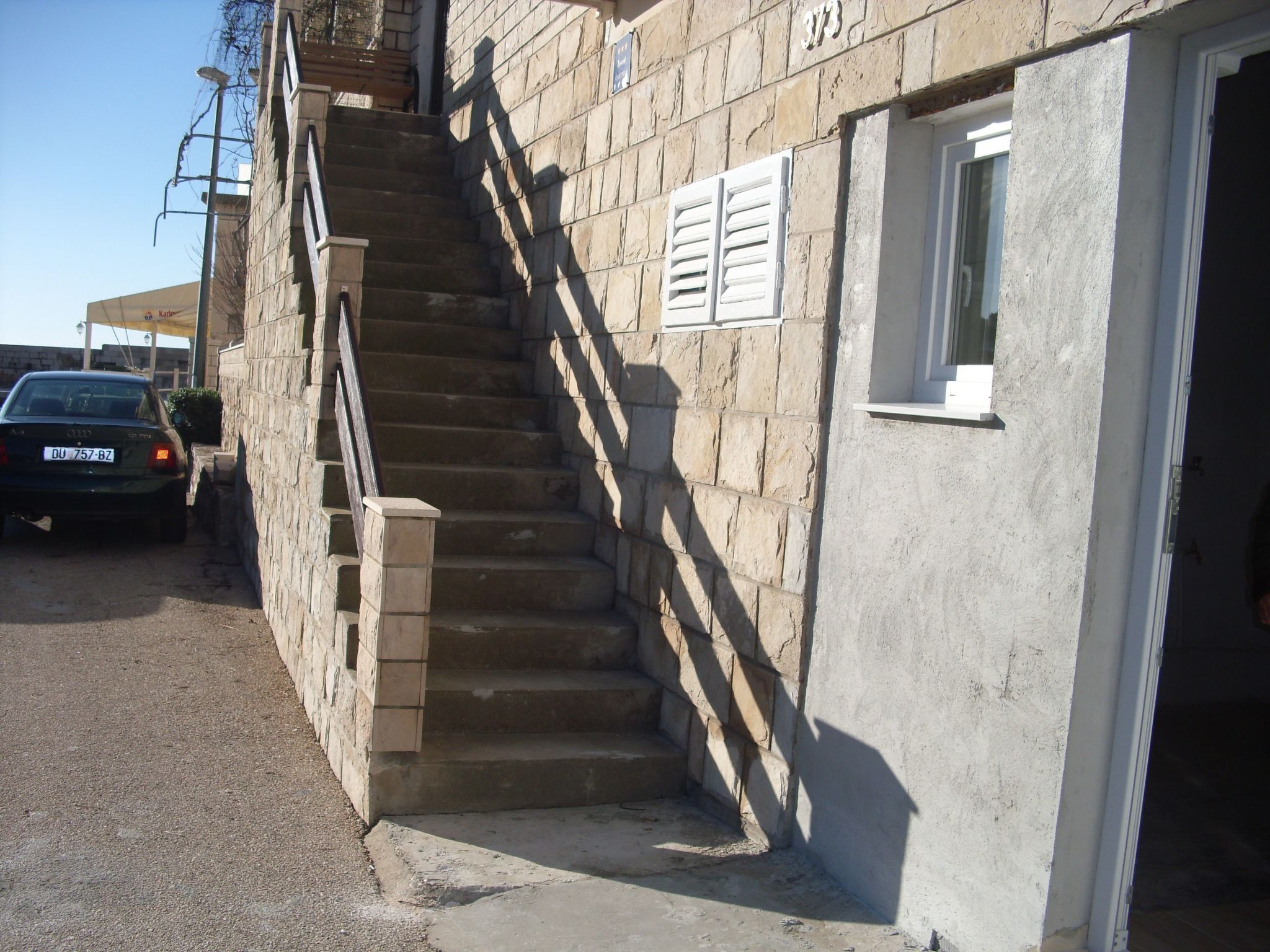 Un escalier en pierre mène à l’entrée d’un immeuble, où une voiture est garée à proximité.