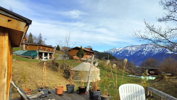 A wooden deck with potted plants and outdoor furniture, overlooking a mountainous landscape with houses and a clear sky.