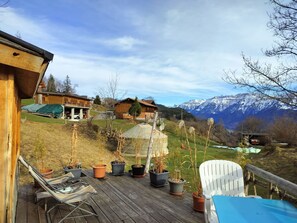 Una terraza de madera con plantas en macetas y mobiliario exterior, con vistas a un paisaje montañoso con casas y un cielo despejado.
