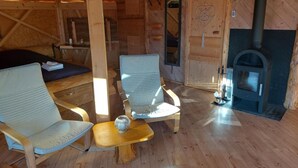 A cozy cabin interior with wooden walls, a fireplace, two cushioned chairs, a small table, and a bed.