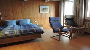Een slaapkamer met een wand van houten panelen, een bed met een kleurrijke deken, twee stoelen en een raam met gordijnen.
