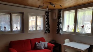 Ein Zimmer mit einem roten Sofa, einem weißen Tisch und schwarzen Stühlen. Es gibt Fenster mit Vorhängen und eine Deckenleuchte.