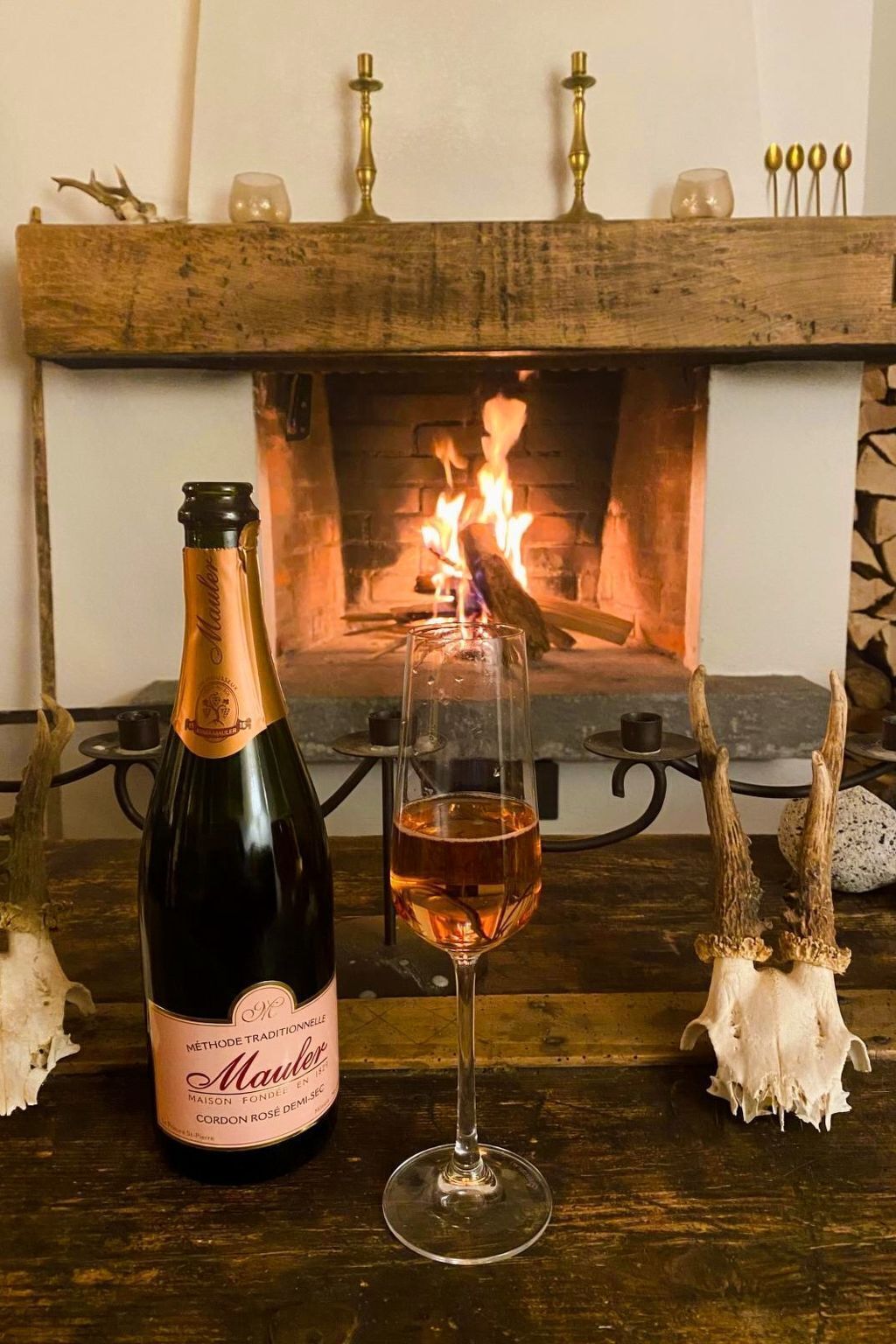 Una botella de vino Maules y una copa de vino frente a una chimenea con fuego encendido.