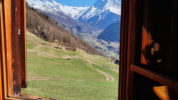 Vista a través de una puerta abierta de un paisaje montañoso con picos nevados, un valle verde y un edificio pequeño a lo lejos.