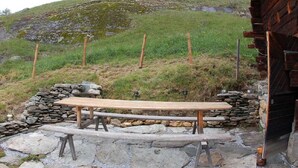 Un área de estar al aire libre rústica con un banco de madera y paredes de piedra, frente a una colina cubierta de hierba y un edificio de madera.