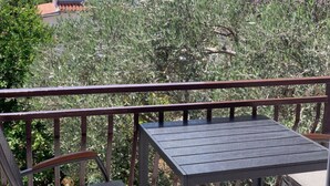 Un balcone con ringhiera in metallo, una zona salotto coperta e vista su vegetazione ed edifici sullo sfondo.