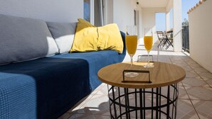 Uma varanda com um sofá azul, um travesseiro amarelo e dois copos de suco de laranja sobre uma mesa de madeira redonda.
