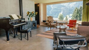 Un salon comprenant un grand piano, un canapé, un fauteuil confortable et une vue sur les montagnes, visible à travers de grandes fenêtres.