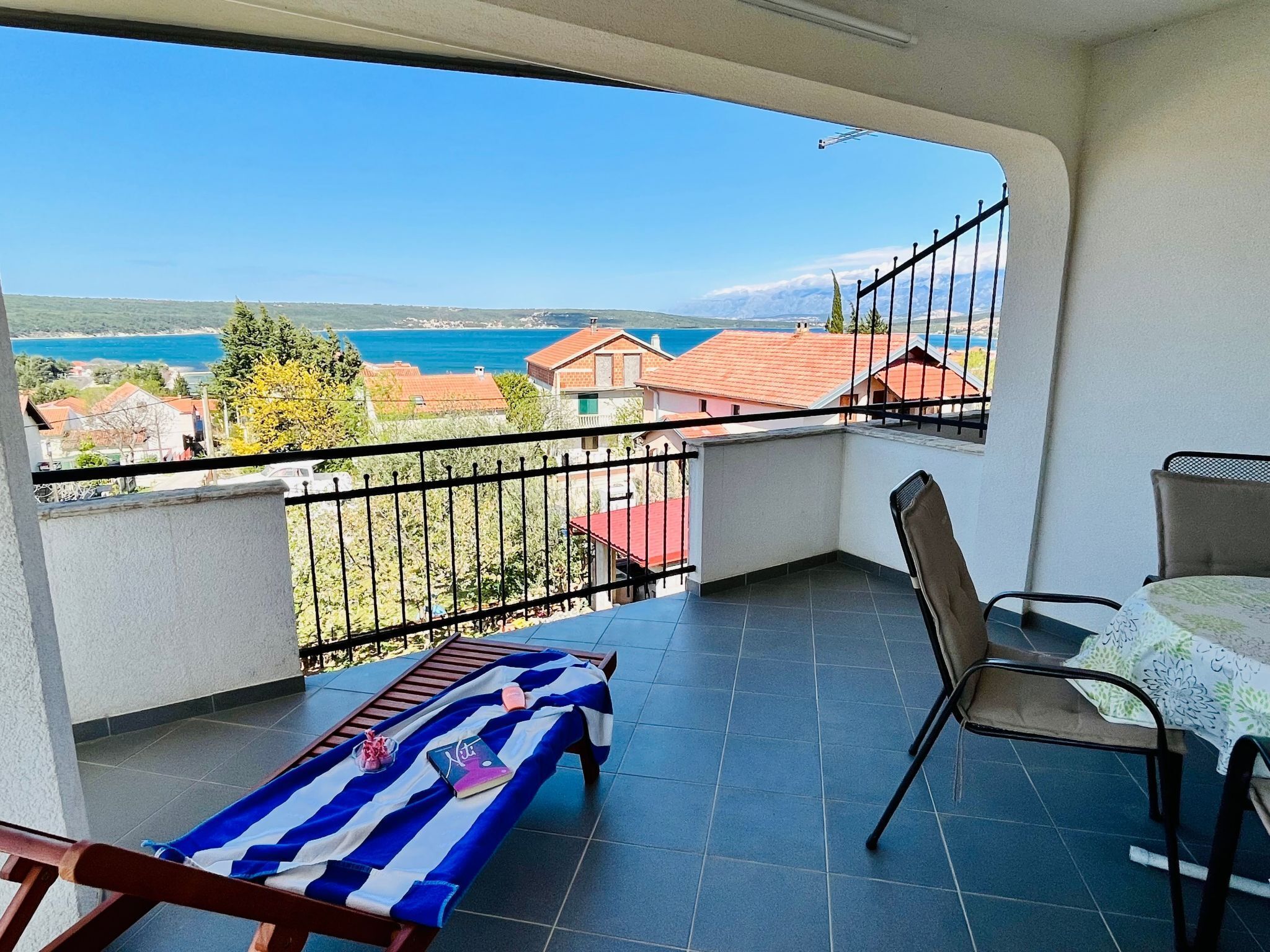 Un balcón con vista al mar y edificios, una mesa con mantel, y una toalla a rayas en un banco.
