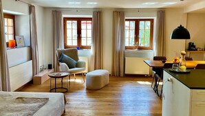 Una habitación de hotel moderna con cama, zona de estar con una silla y una mesita, kitchenette y ventanas grandes con cortinas.
