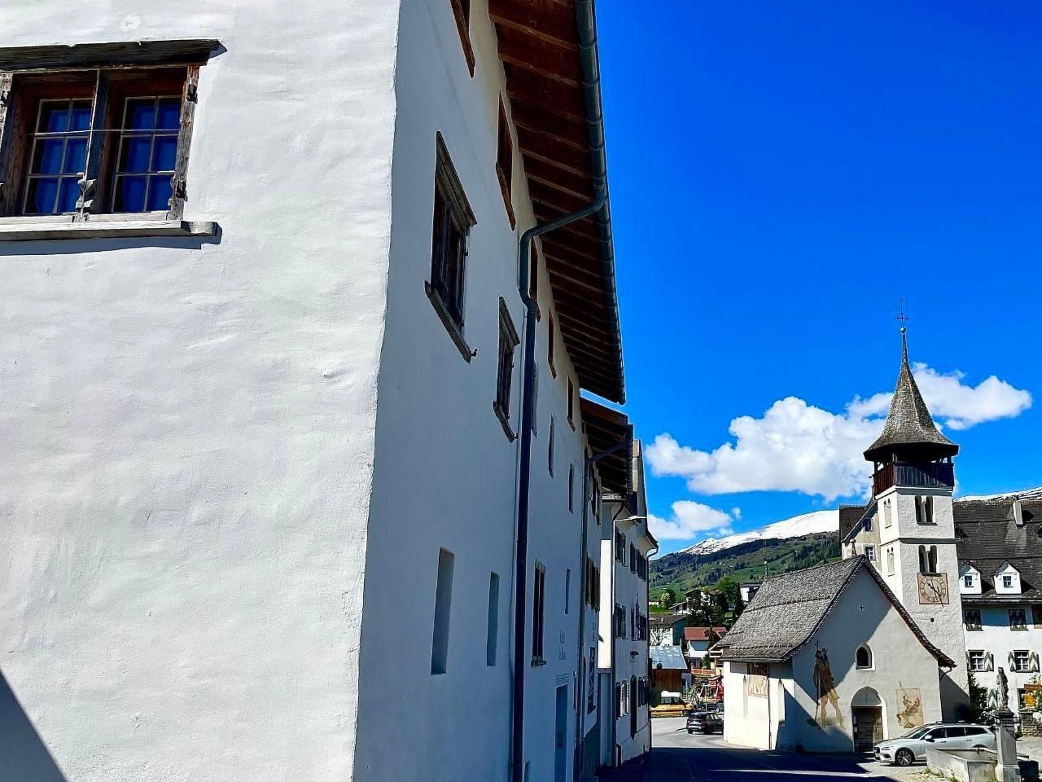 Una calle estrecha con edificios blancos, una iglesia con un campanario puntiagudo y una montaña al fondo.