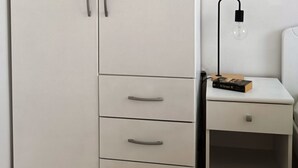 Un armadio bianco con due ante e tre cassetti, un comodino con una lampada e una piccola mensola.