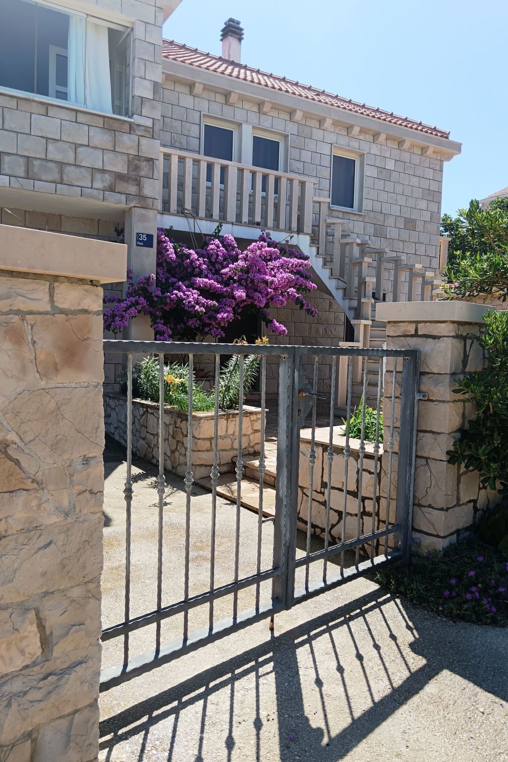 Un edificio de piedra con techo de tejas, entrada con portón y una caja de flores con flores moradas.