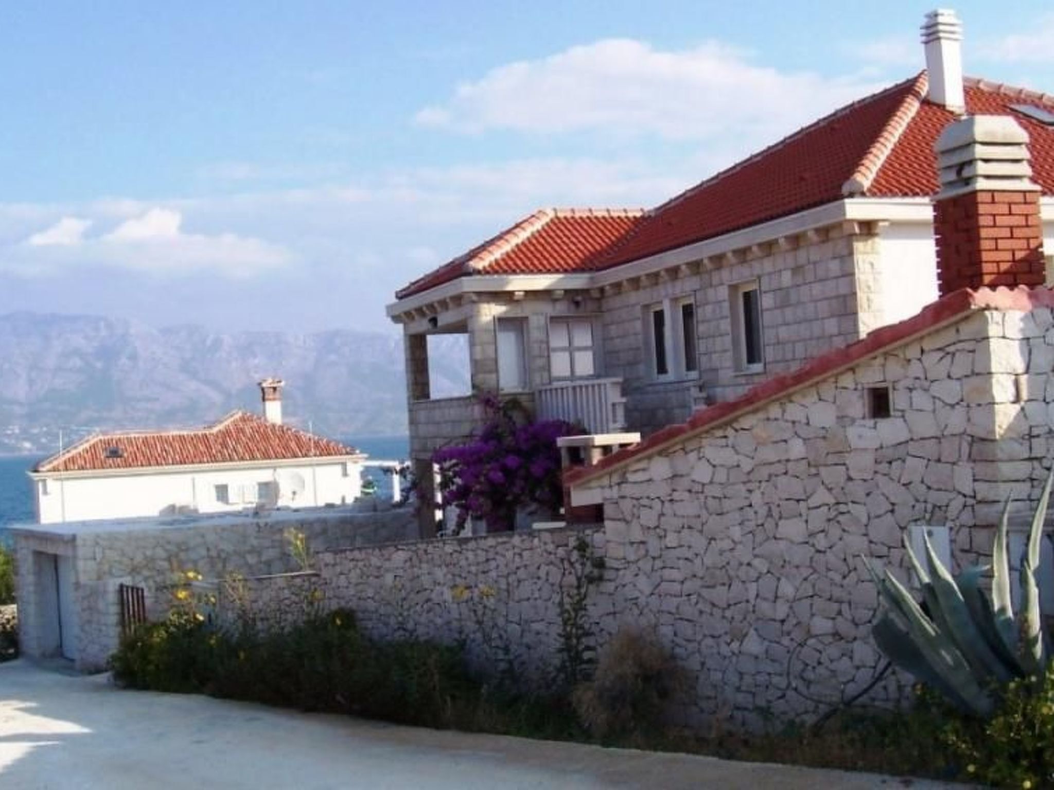 A stone building with a red-tiled roof, a balcony, and a view of the sea and mountains in the background.