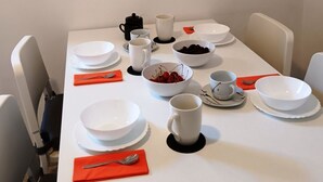 Una mesa de comedor dispuesta con platos, tazones y tazas blancos, junto con servilletas rojas y una cuchara.