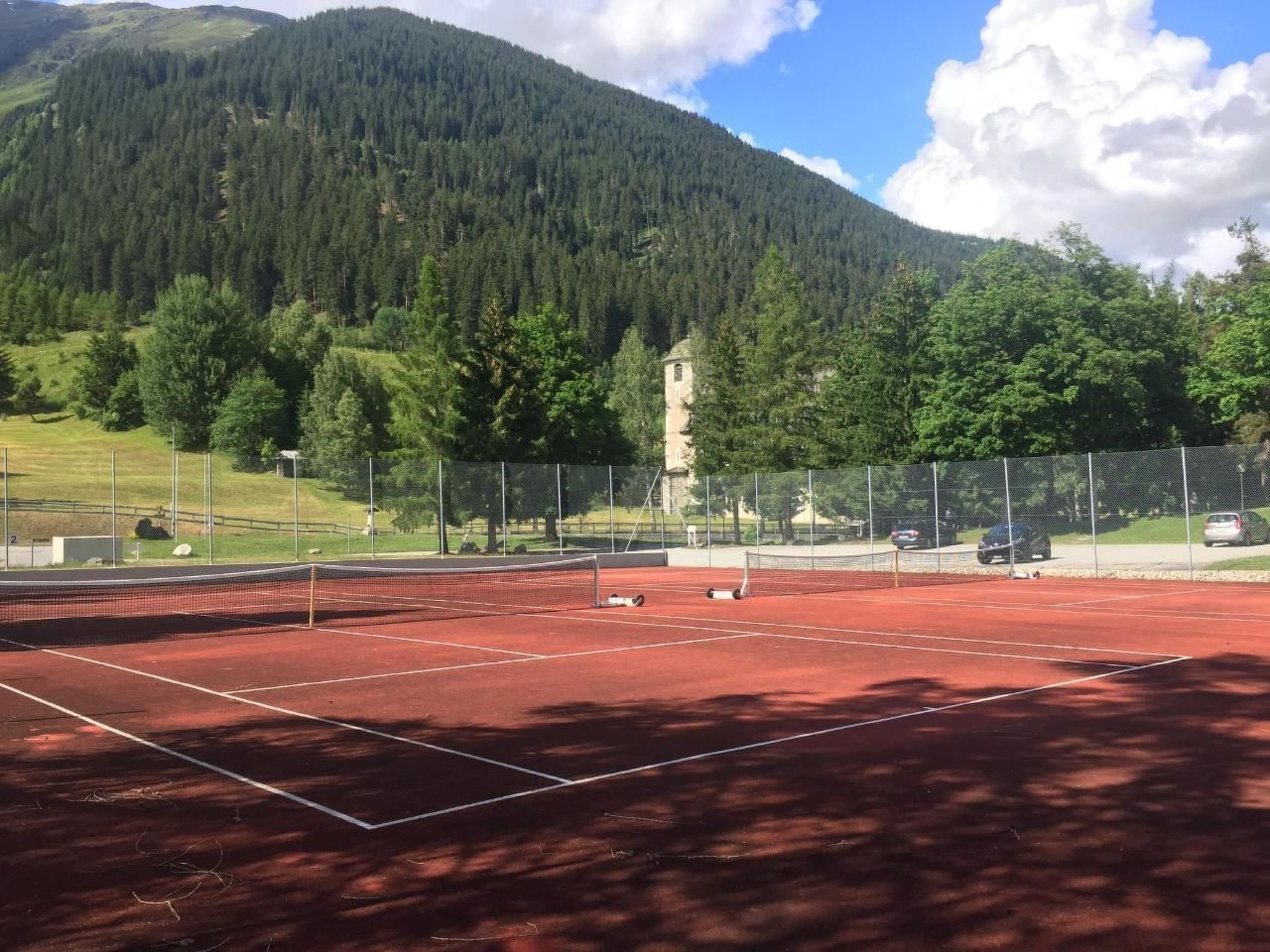 Een tennisbaan met een net, omgeven door een hek, met op de achtergrond een bebost gebergte en een heldere lucht.