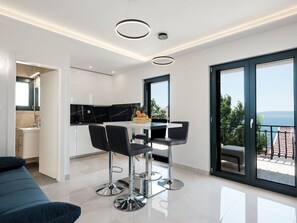 Eine moderne Küche mit weißen Schränken und einer schwarzen Marmor-Spritzschutzwand, ein Essbereich mit weißem Tisch und schwarzen Stühlen sowie ein Balkon mit Blick auf das Meer.